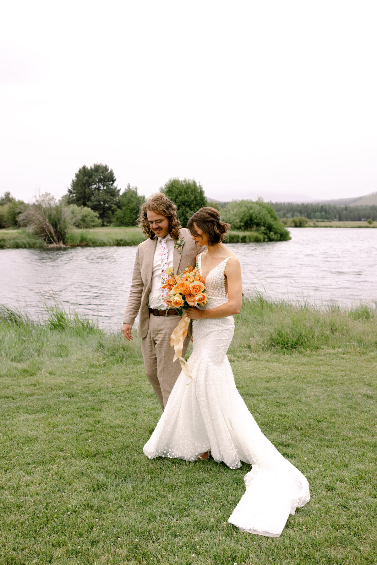 Young wedding couple walking in front of lake view holding florals and smiling representing full service design and calligraphy by Life in Letters.