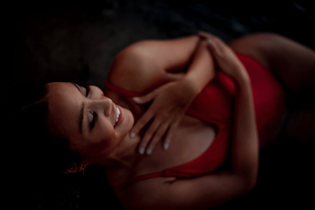 woman in a red bathing suit smiles and embraces herself during her beach boudoir session