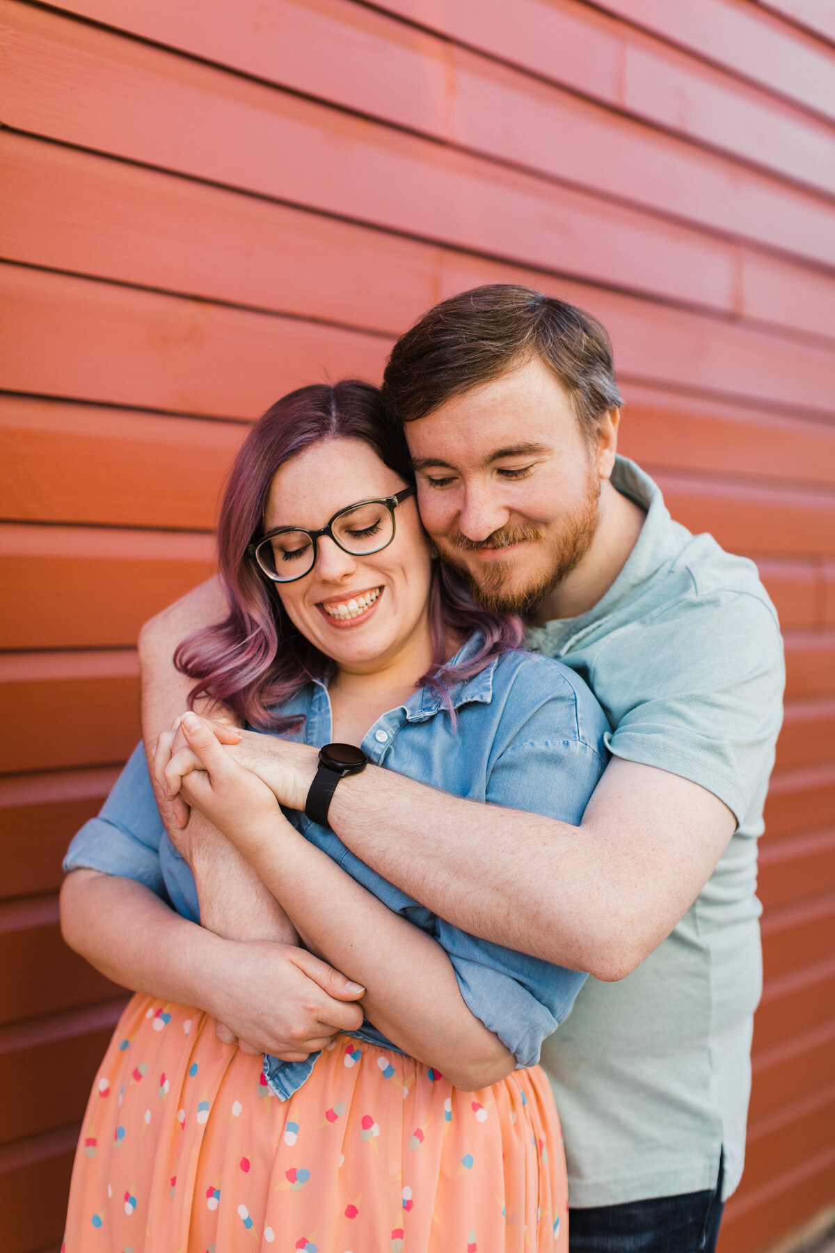 A couple acting as the big spoon and little spoon in front of a red wall during their engagement session in Fort Worth, Texas. The woman on the left is the little spoon and is wearing a jean jacket and a salmon skirt with colorful polka dots and glasses. The man on the right is the big spoon and is wearing a short sleeve collared shirt and jeans.
