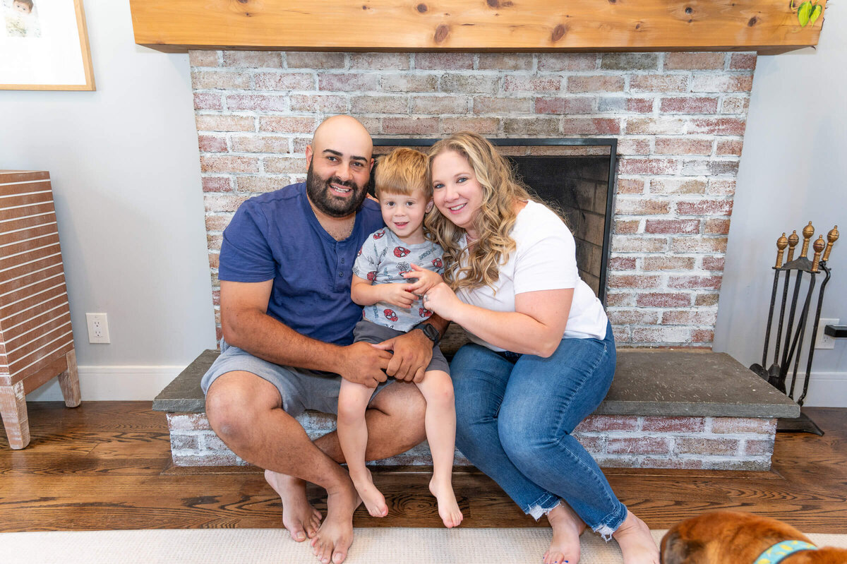 Parents with their toddler son, sitting on their fireplace, close together, smiling at their home in Greenwich, CT.