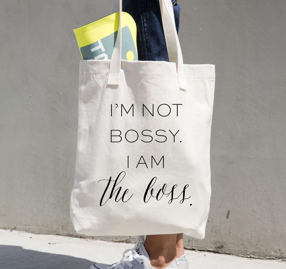 A person carries a beige tote bag with the text "I'M NOT BOSSY. I AM the boss." printed on it; a yellow item is partially visible in the bag, showcasing Sweet Pea Ink Creative’s bold approach to brand design.
