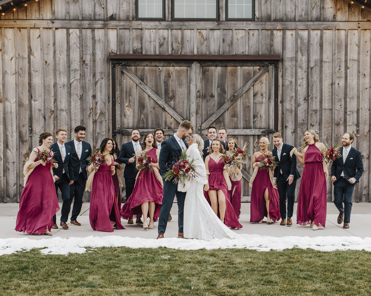 Joyful wedding party celebrating outside a rustic barn with bridesmaids in deep red dresses and groomsmen in suits, while the bride and groom share a kiss in the foreground taken by jen Jarmuzek photography a Minneapolis wedding photographer