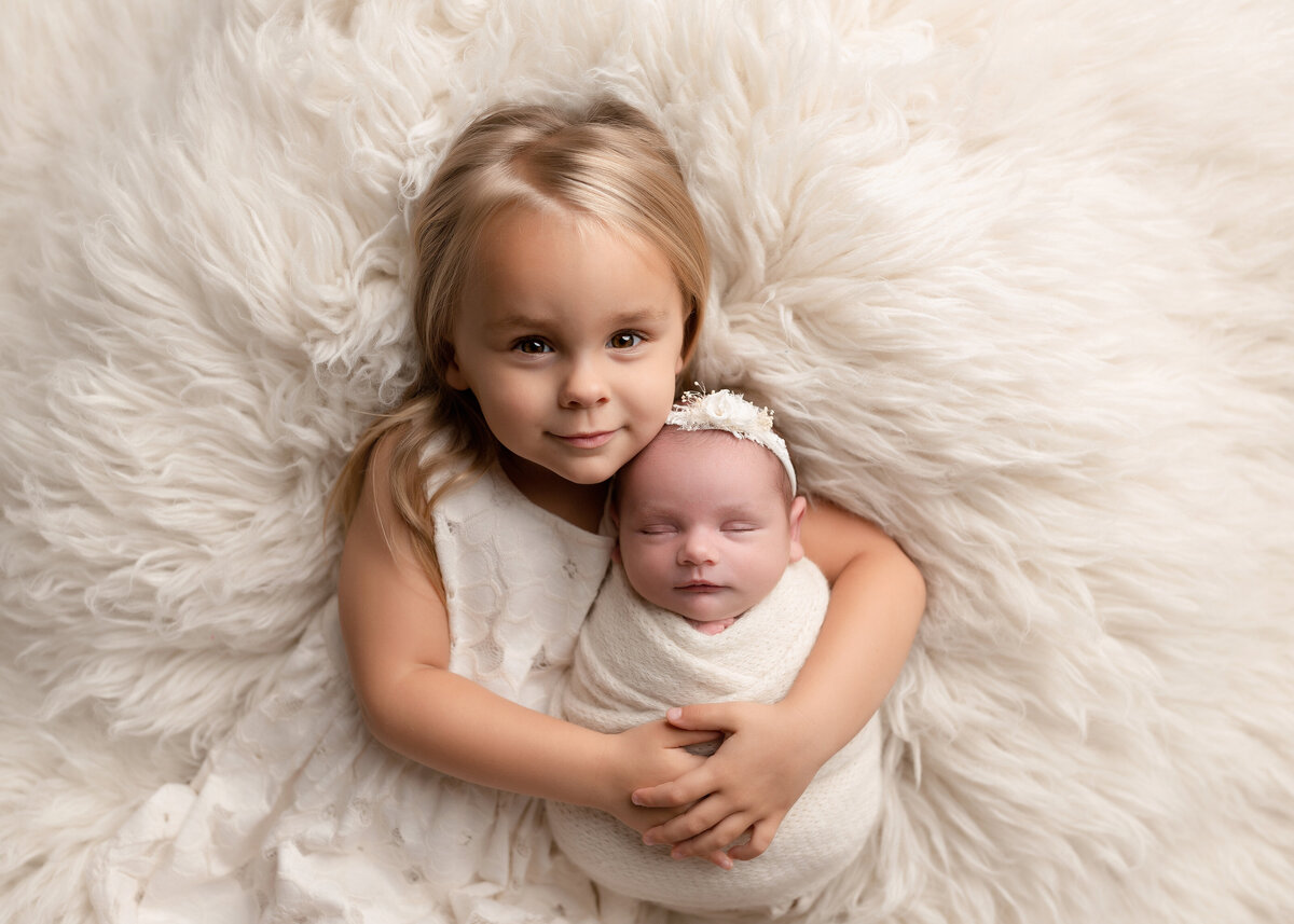 Newborn photoshoot in Delray Beach and West Palm Beach photography studio - sibling photo. Big sister has her arms wrapped around baby sister atop a cream colored faux fur rug. Big sister is smiling at the camera, baby sleeping with her head resting on big sister's shoulder. Baby is wearing a delicate floral headband. Aerial image.