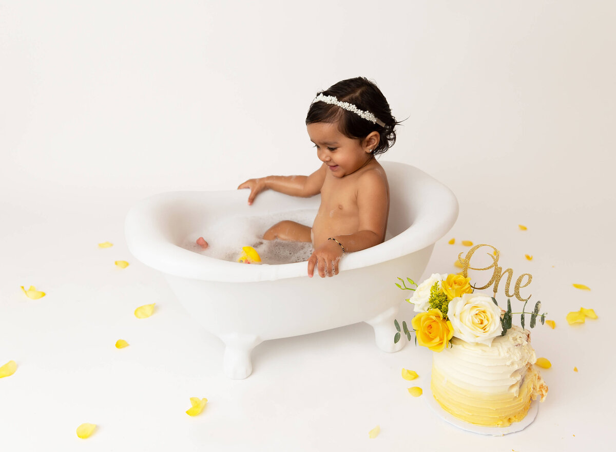 Baby girl sits in a bathtub to clean up from cake smash photoshoot. In front of the tup there is the smashed yellow ombre cake  during Cake smash photoshoot in NYC 
