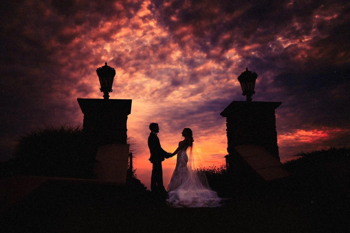 Bride and groom holding hands in silhouette against a dramatic red and purple sunset sky, between two ornate lamp posts