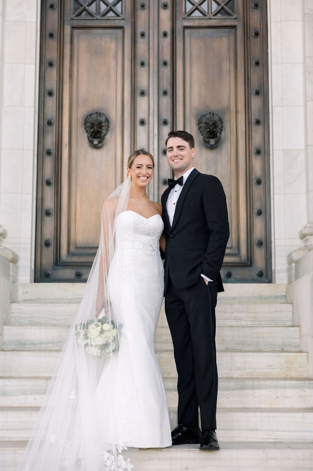 A bride in a white dress and veil holds a bouquet, standing next to a groom in a black tuxedo. They are smiling in front of an ornate wooden door with lion head embellishments.