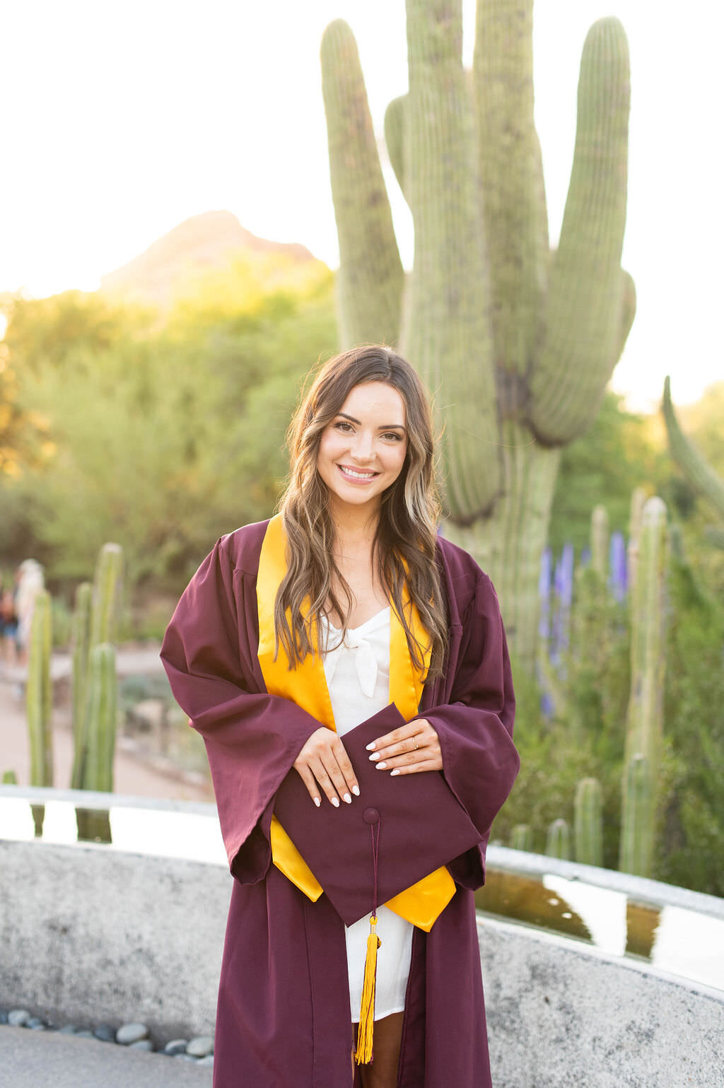 A woman smiling while holding her graduation cap in a small garden.