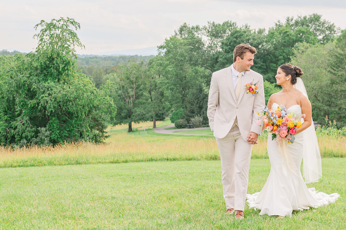 A stunning couple in a light cream colors and a bright wedding bouquet holding hands and walking in the Virginia mountains in June  