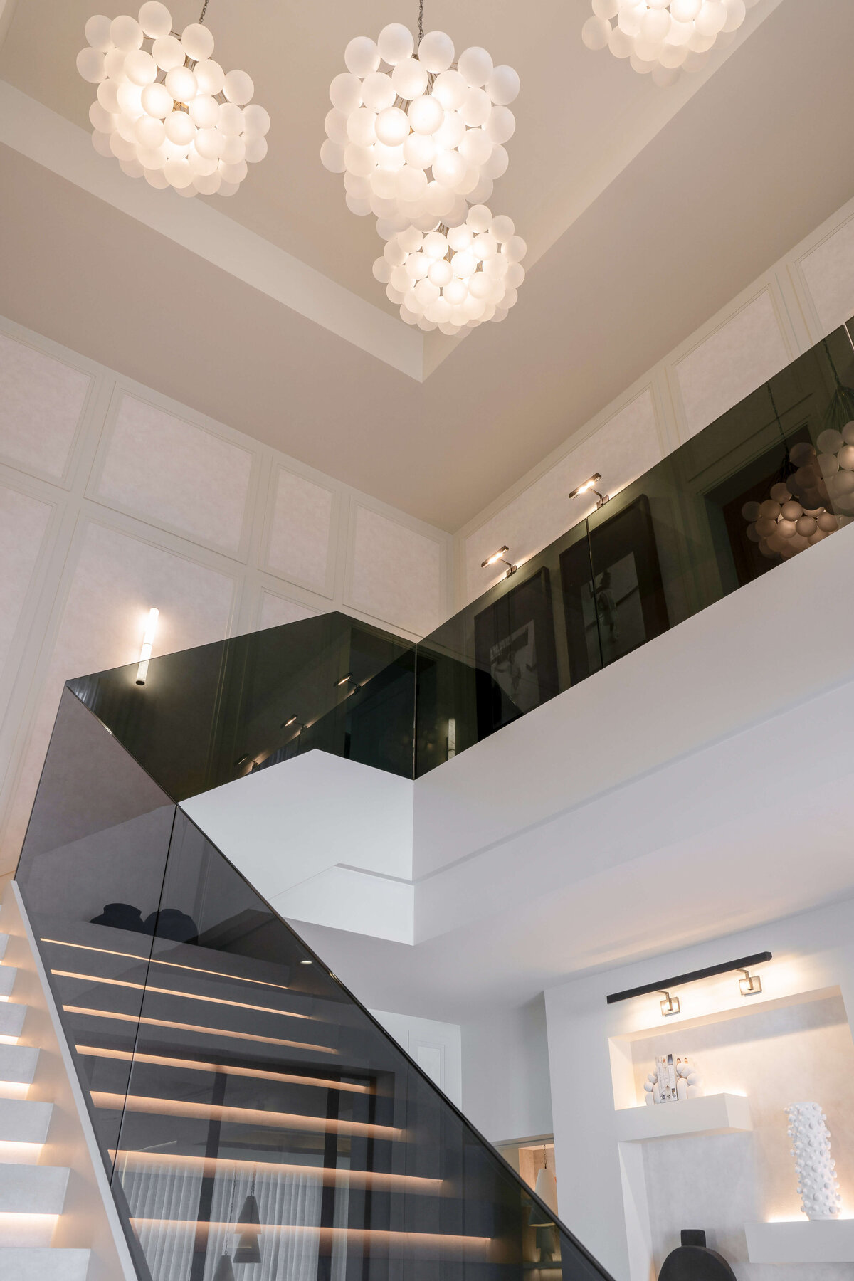 The top of a modern white staircase, bordered by a sleek, black glass railing on the right side. The staircase leads to a large, open landing, with the black glass railing continued through the hallway. Above, bold, bubbly chandeliers emit a soft light and add a contemporary style to the space.