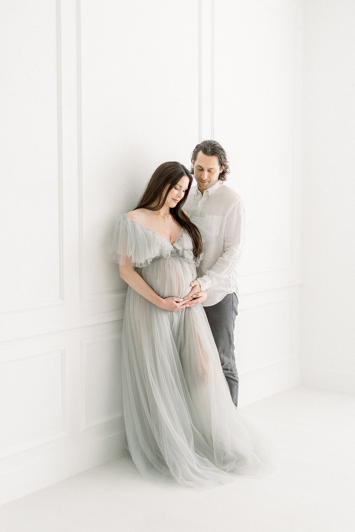 Portrait of an expecting Dallas mom leaning on a wall with her spouse standing in front of her as he holder her belly for their maternity session at a Dallas photography studio.