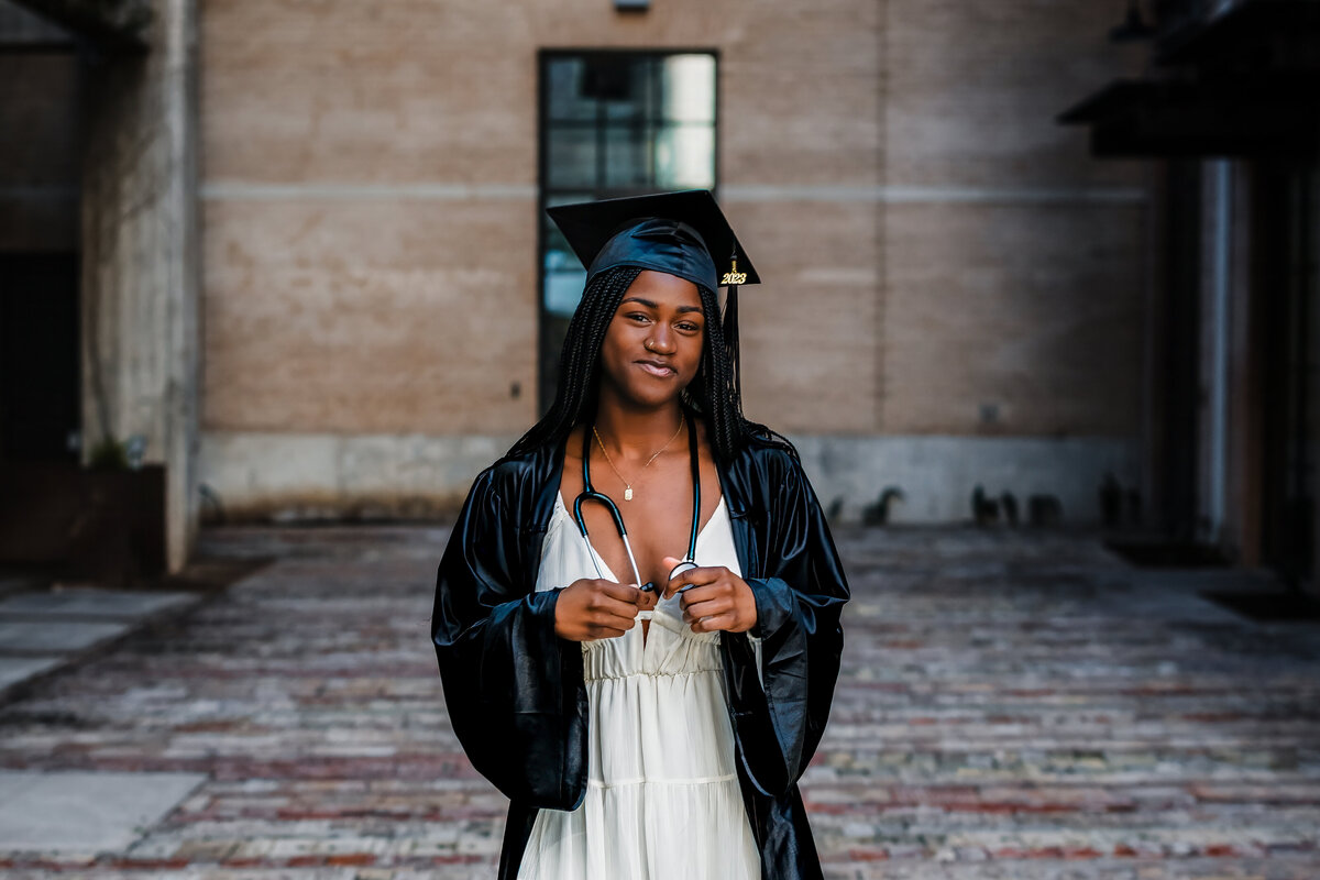 Steel High School senior girl holding stethoscope while wearing white dress and black gown at The Pearl.