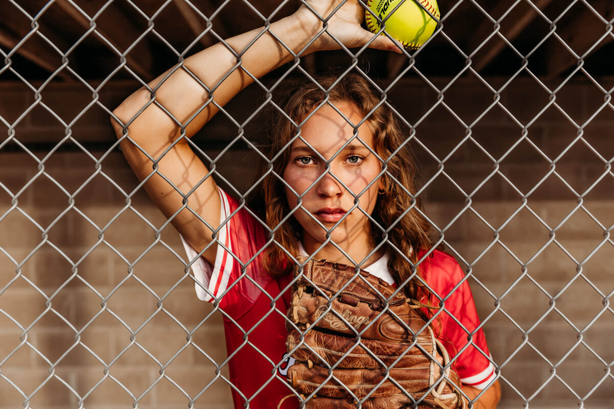 gorgeous senior leaning against the fence in softball dugout with softball