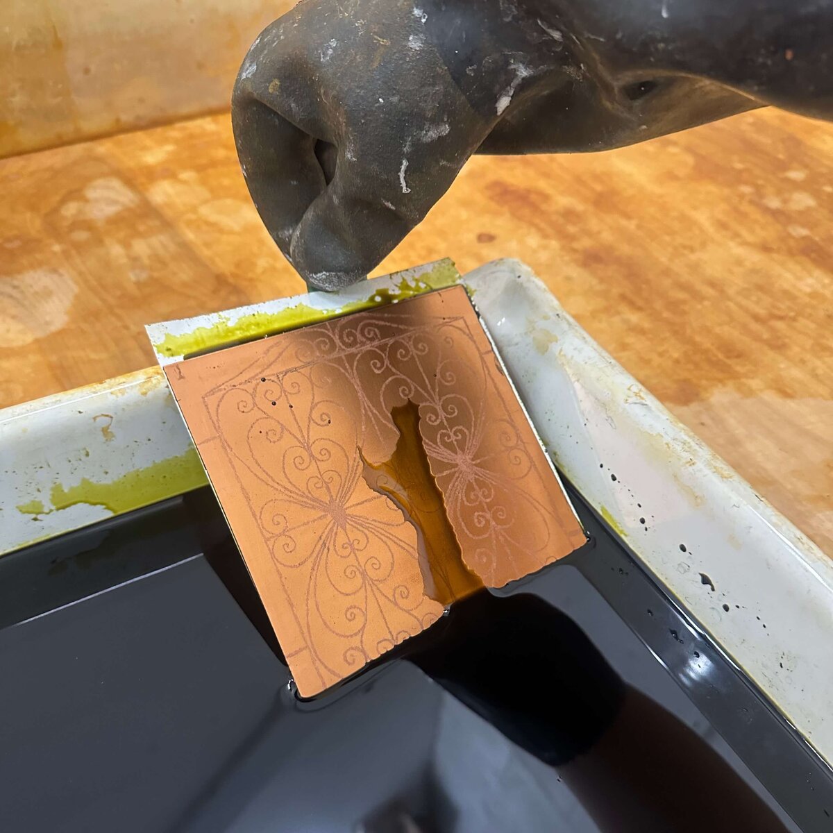 A gloved hand lowers a copper plate into an acid bath mid-etch, revealing the etched design as it develops.