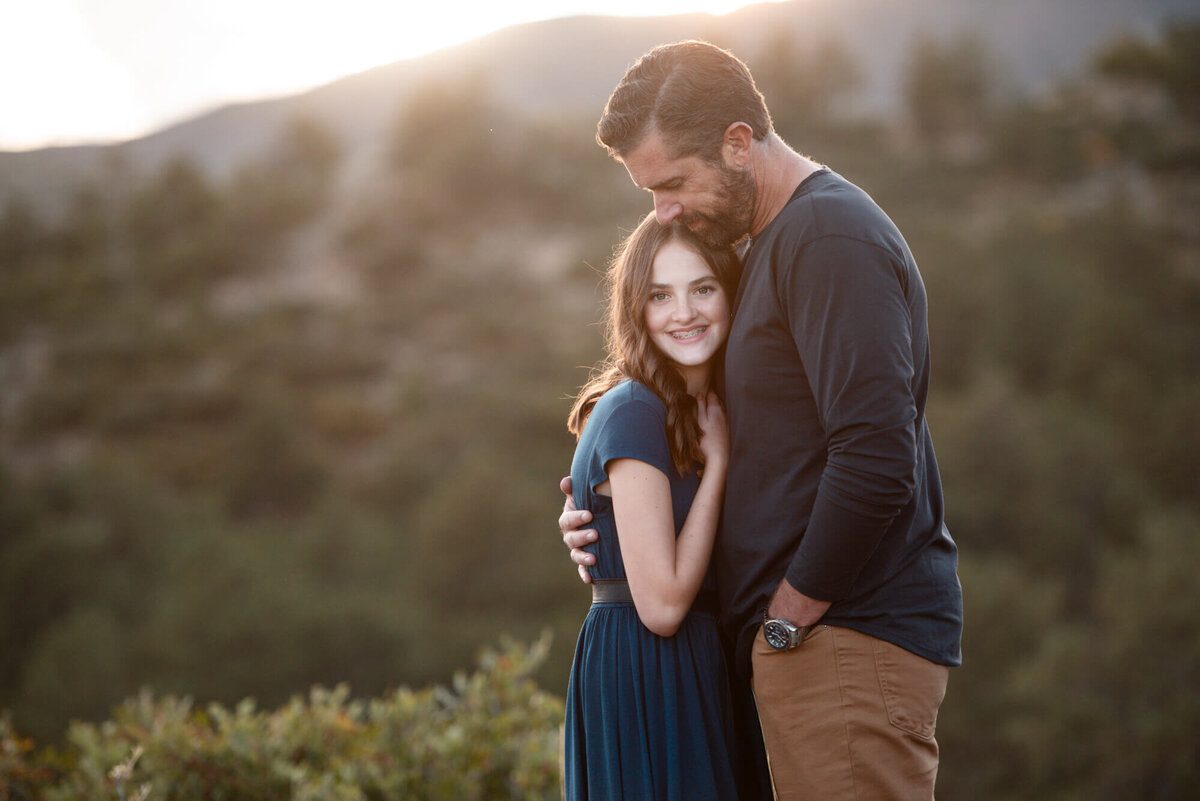 A father hugs and snuggles his teen daughter in a blue dress while standing outside at sunset