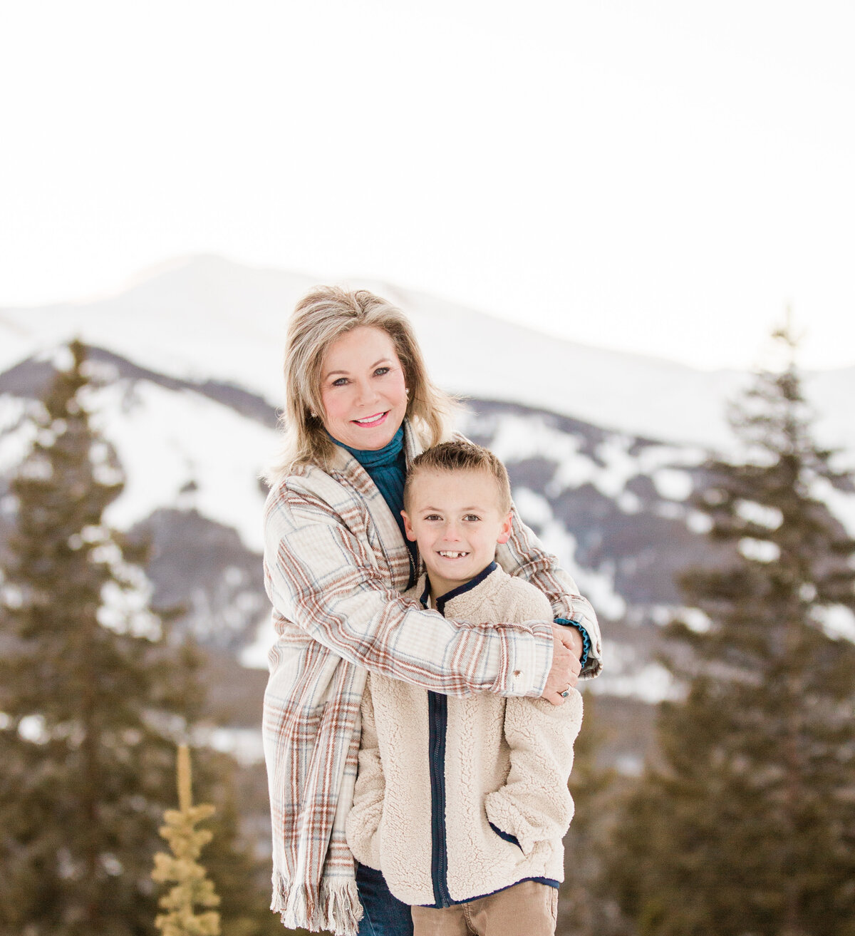 A grandma is hugging her 8 year old grandson. It is winter and the snowy ski runs of Breckenridge Resort are behind them