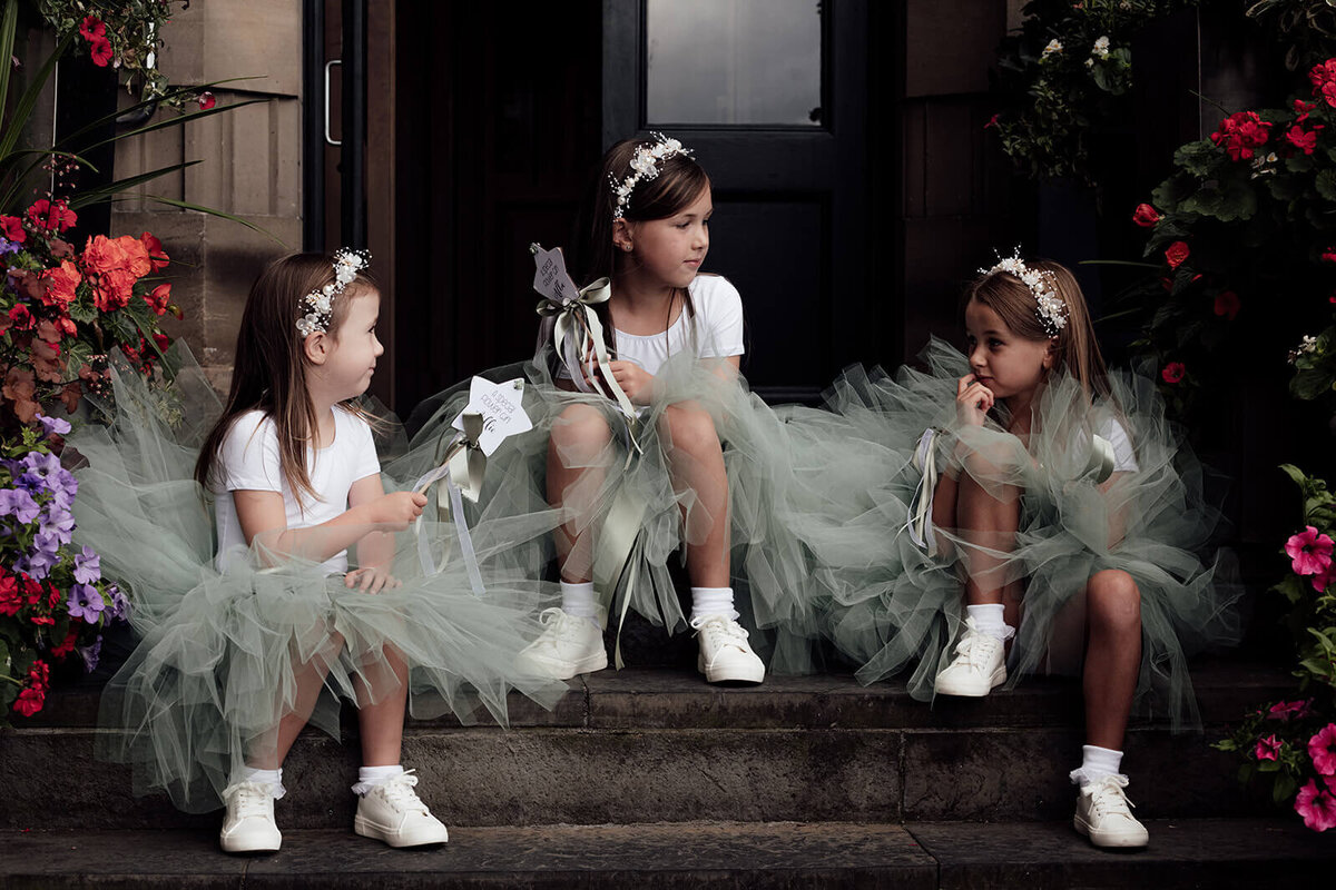 Three young bridesmaid's wave their wands as they sit and play. They are wearing green tulle dresses. 