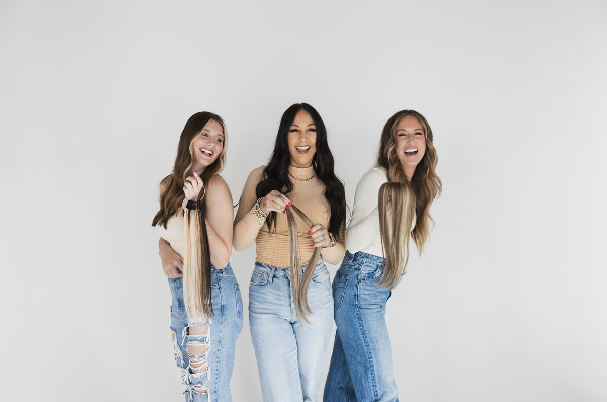 Three hair extensions models are standing together and smiling while holding hand-tied wefts and a color swatch ring.