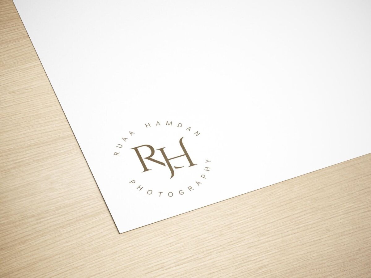 lose-up of a white sheet of paper on a light wood surface, featuring a minimalist circular logo reading “RH Ruaa Hamdan Photography” in elegant serif lettering.