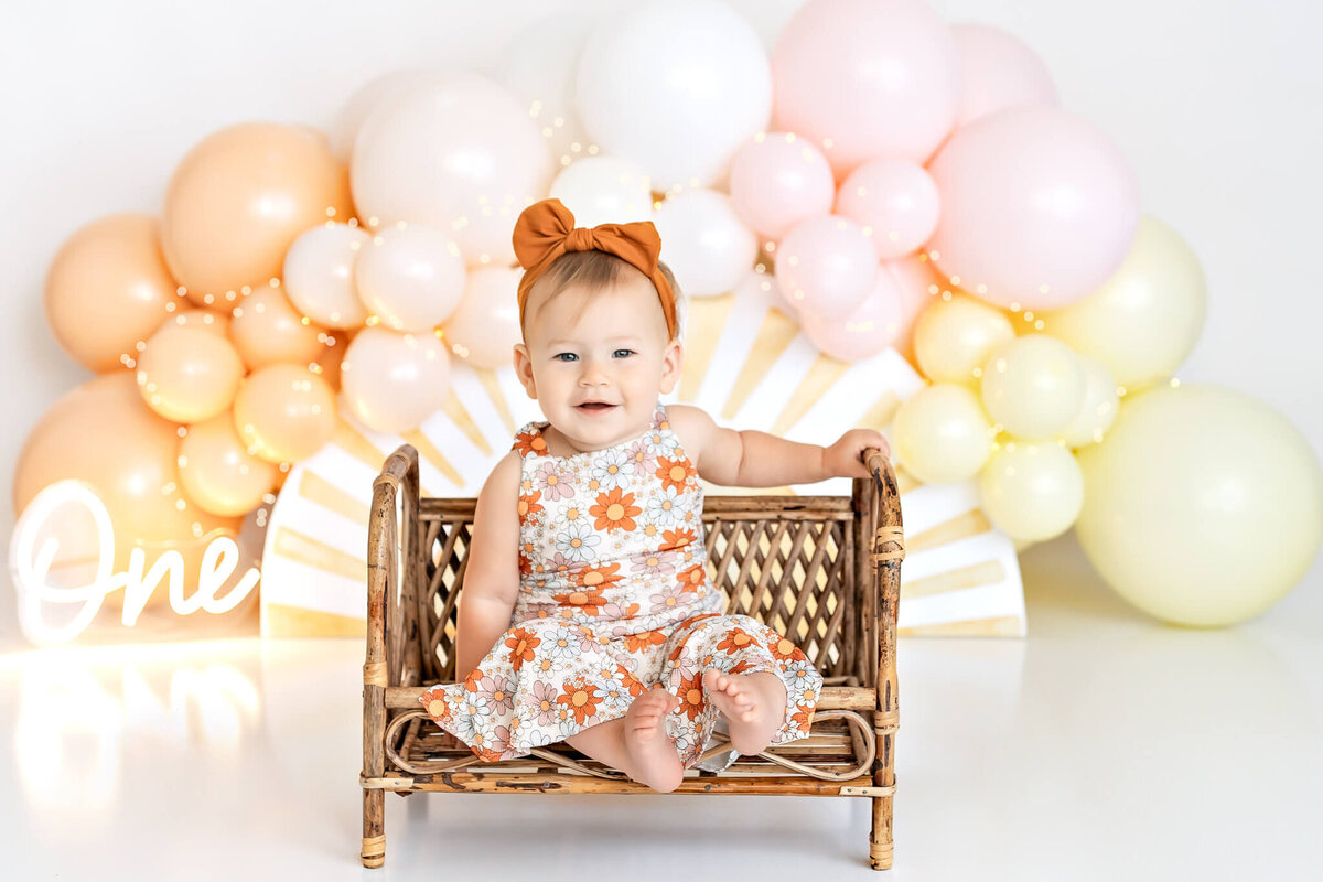 A baby in a floral dress and headband sits on a wicker chair. Behind her, a pastel balloon arch in shades of peach, pink, and yellow with a "One" sign is visible, suggesting a first birthday celebration.