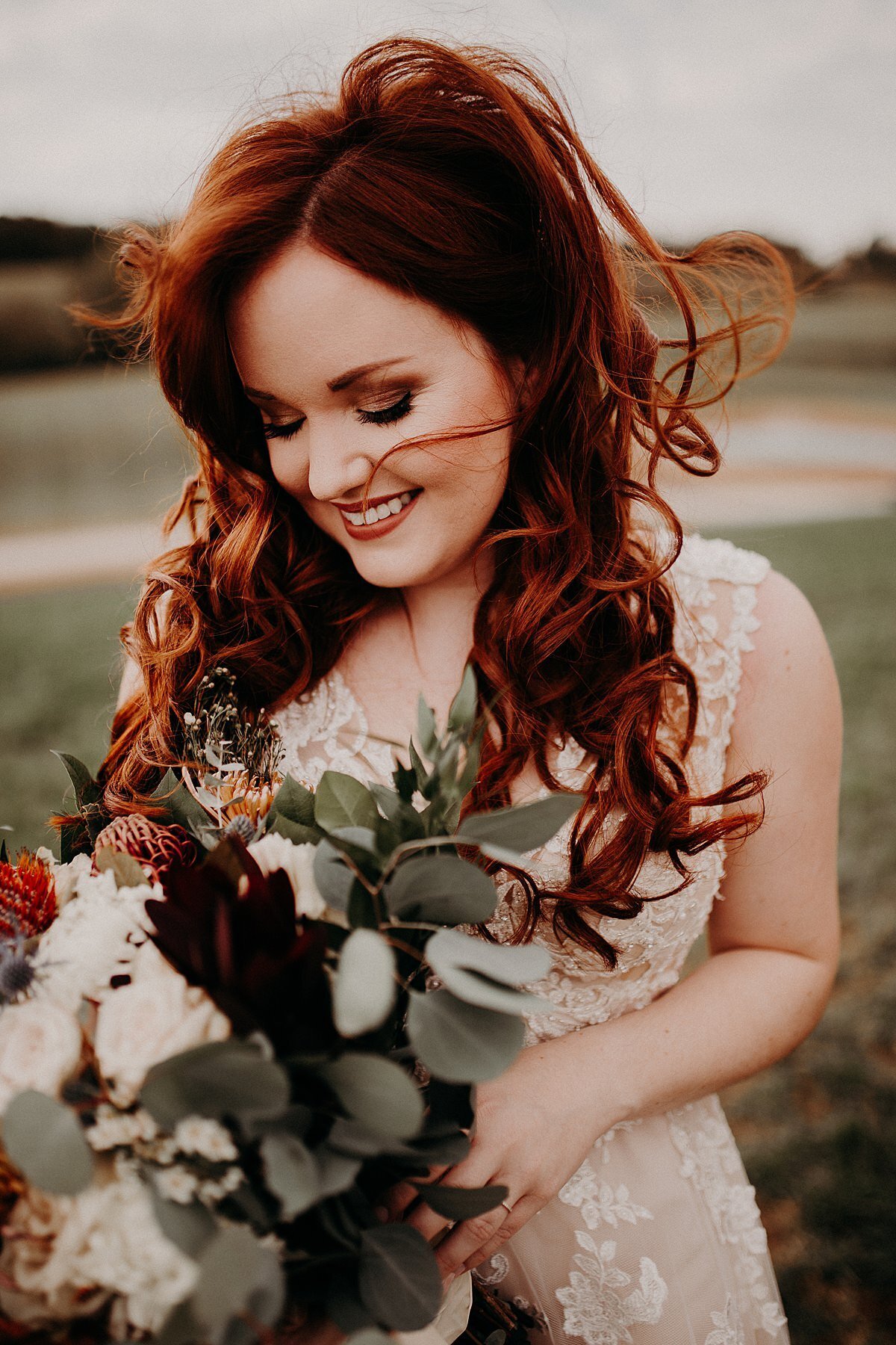 Red haired bride smiles down at her dark burgundy, ivory and rust colored bridal bouquet wearing a beaded lace wedding dress as the wind blows around her.