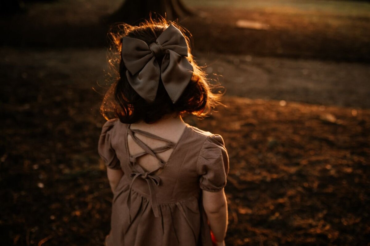 a little girl with a prominent bow in her hair, standing in a park at sunset with golden light illuminating her hair and dress.