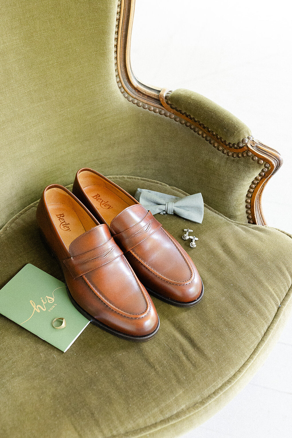 Souliers brun du marié, son carnet de voeux et son noeud papillon sur un fauteuil vert, mariage au Manoir Maplewood.