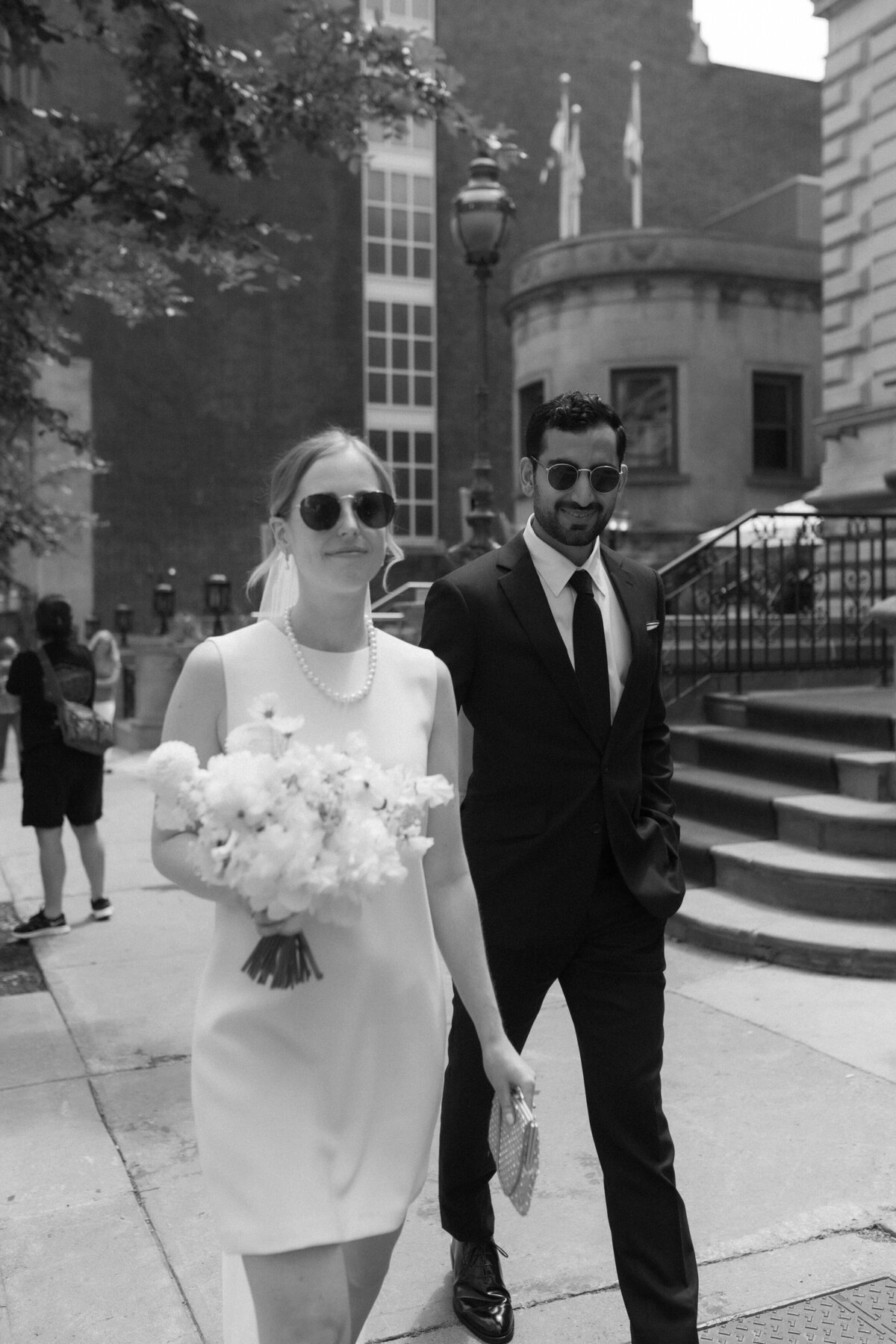 A couple wearing sunglasses walk down a street in old Montreal, the bride holds a bouquet.