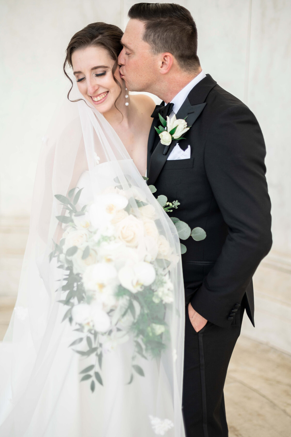A groom in a black tuxedo gently kisses a smiling bride on the cheek. The bride, in a white gown with a sheer veil, holds a lush bouquet of white and green flowers. The setting appears elegant and bright.