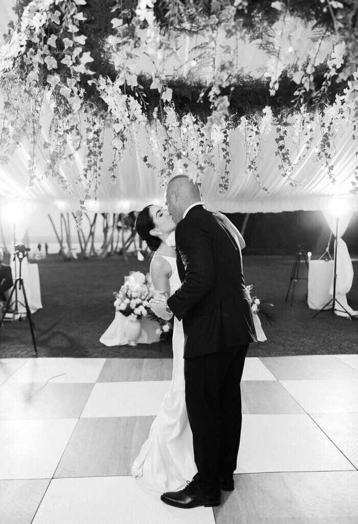 An intimate black-and-white portrait of the newlyweds sharing a kiss on the dance floor, surrounded by lush floral arrangements and glowing lights. Captured by Claudia Amalia Photography.