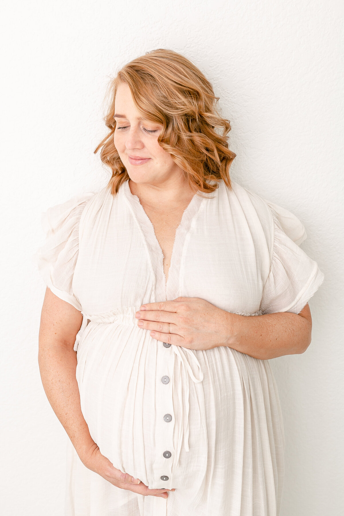 Pregnant Woman dressed in white holding belly and looking down at belly during Studio maternity session in Portland Oregon.