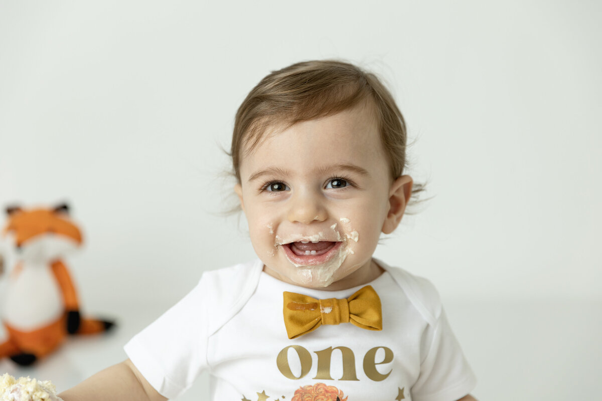 A happy toddler boy in a gold bowtie smiles big while covered in cake frosting