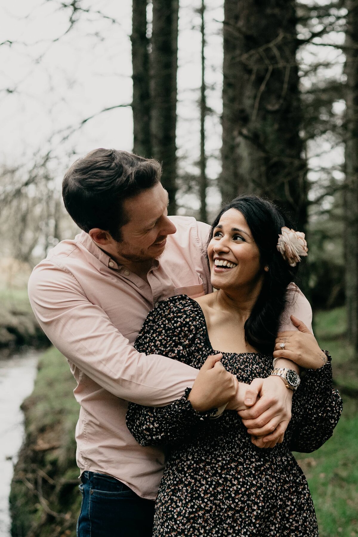 A man stands behind his fiancé, wraps his arms around her and they smile candidly at each other during their engagement photoshoot.