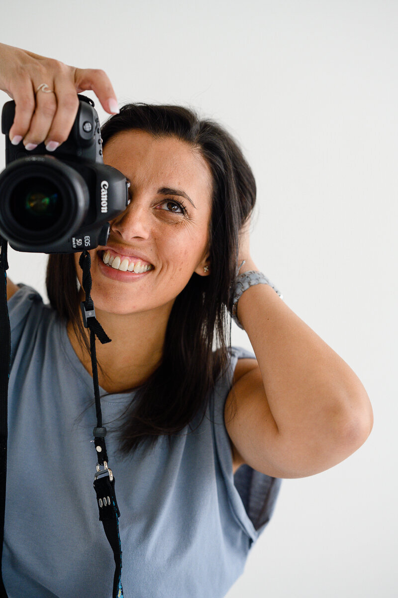 A denver woman photographer poses for a denver commercial photo shoot of her holding her camera up to her face while wearing a blue shirt and smiling for her branding photos and branding pictures.
