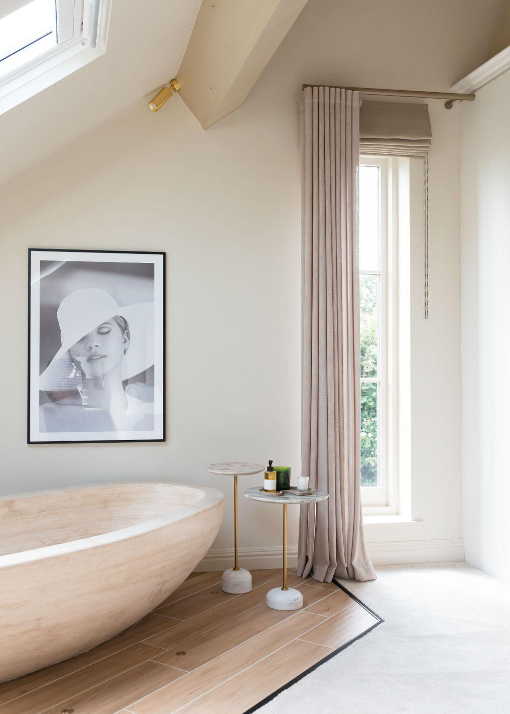 A minimalist neutral bedroom features a large, freestanding wooden bathtub under a skylight. There's a small side table with decor and a framed black-and-white photo on the wall. The space is bright with natural light and neutral tones.