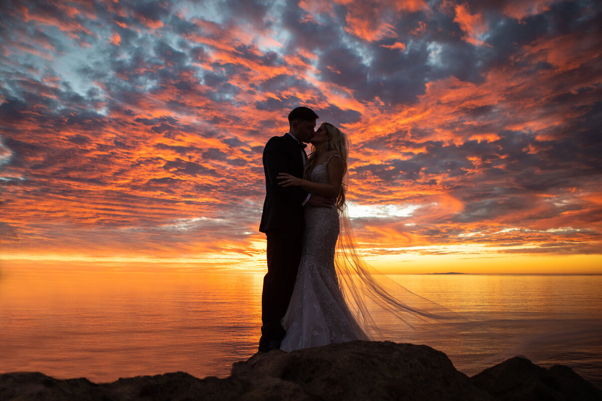 bride and groom kiss on hilltop with colorful sunset and ocean behind them