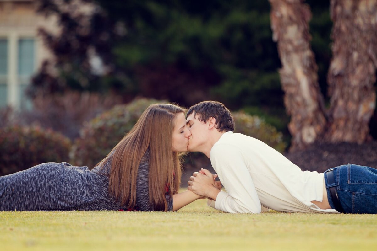 Couple lying in the grass on their stomachs facing each other for a kiss for their engagement portrait.