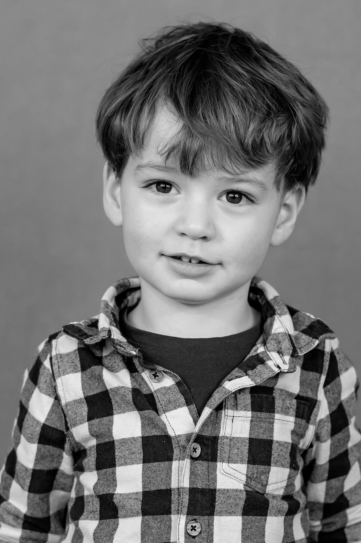 Preschool boy wearing a plaid button down shirt smiles sweetly for his portrait
