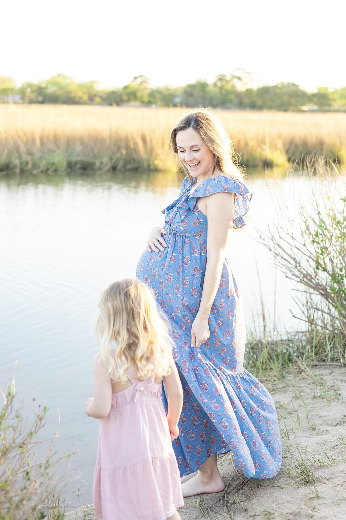A pregnant mother wearing a blue floral pattern dress stands on the shore with her young daughter by  Maternity Photographer Virginia Beach