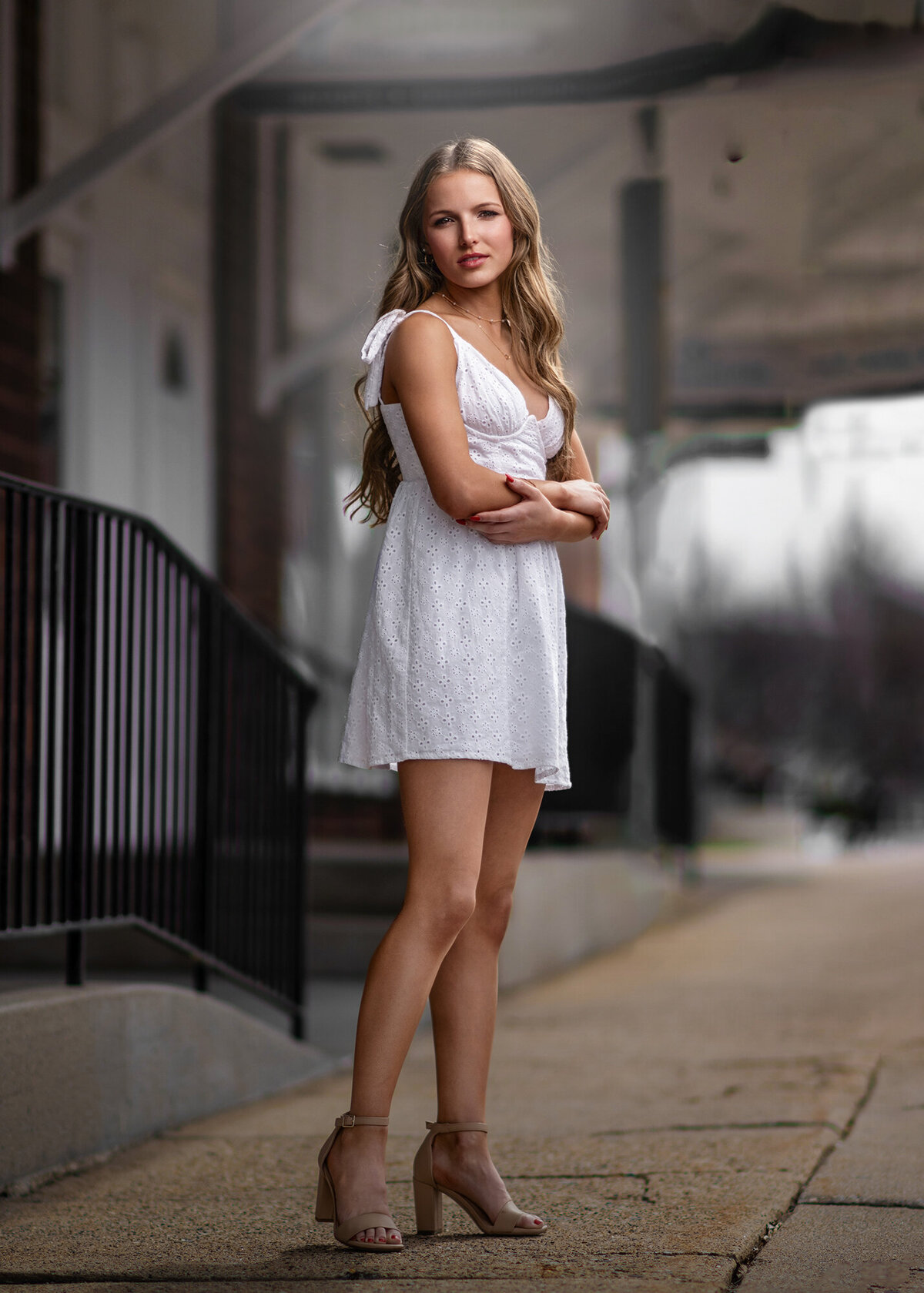 A serious Pella senior in a white dress stands on a sidewalk with her arms crossed