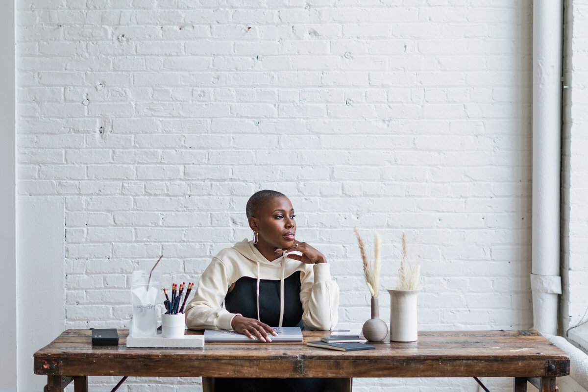 An editorial-style wide-angle image of the artist in an ivory and black outfit, seated at her desk and gazing out the window. The composition blends minimalist design with a sense of calm, creative reflection.