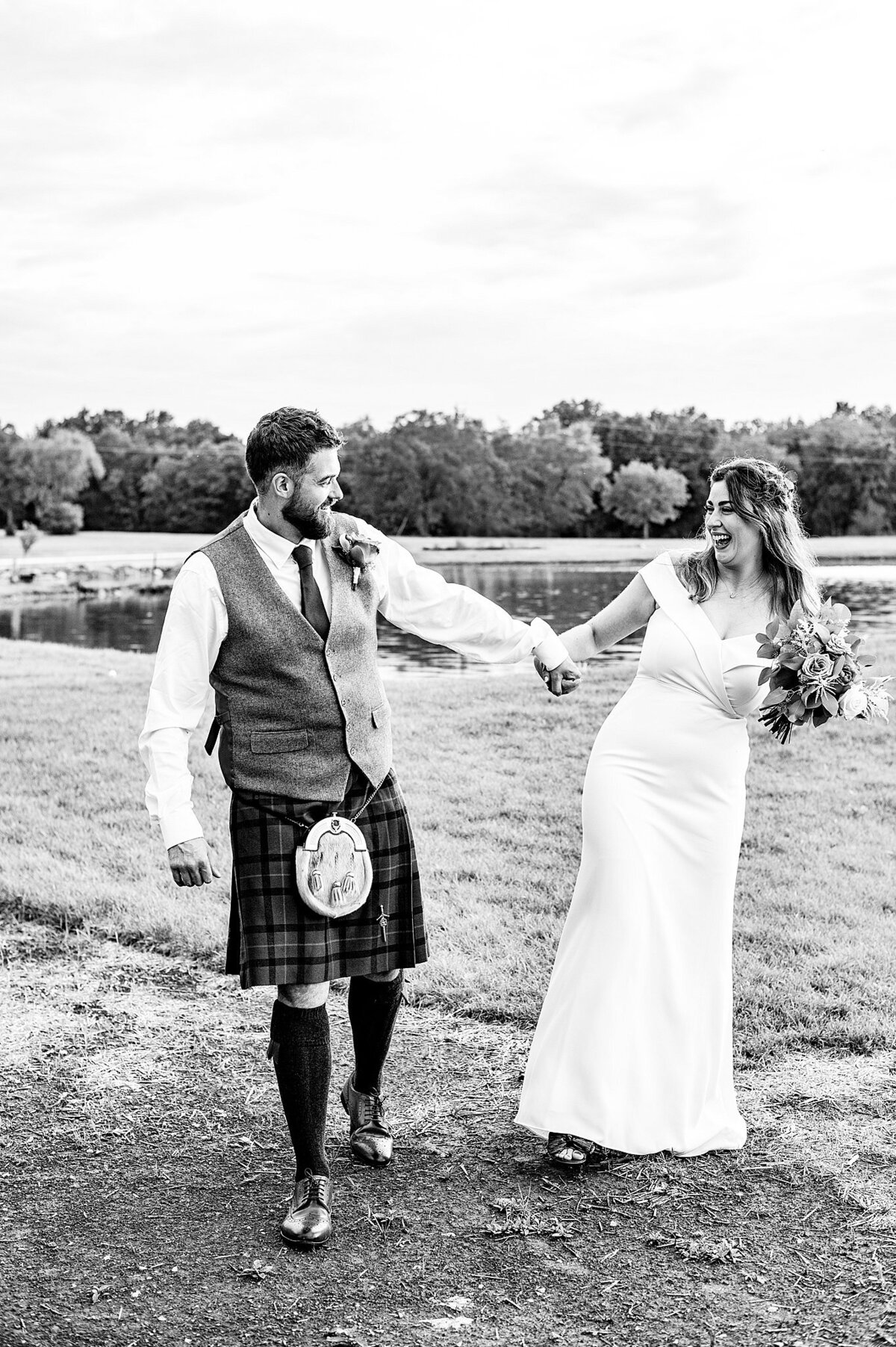 The groom, wearing a traditional Scottish kilt and sporran dances hand in hand with the bride who is laughing and twirling while holding her bouquet beside a lake at their Nashville elopement