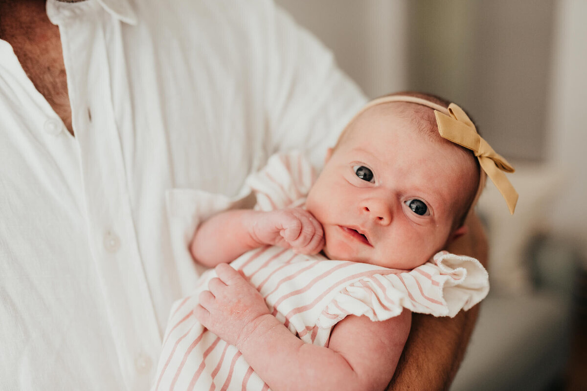 A newborn baby girl held by her father and wearing a white and pink striped outfit and golden bow looks at the camera during her photoshoot with Allison Wolf, Boston Newborn Photographer
