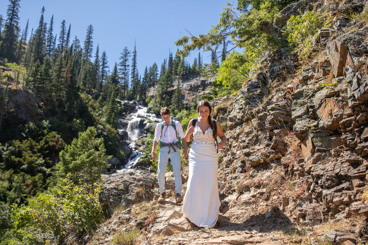A bride and groom wearing backpacks, hike down a rocky trail with a waterfall flowing behind them.