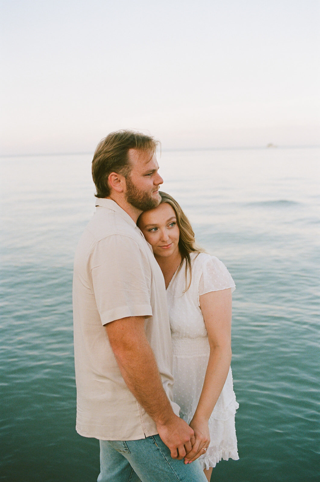 A couple stands by the water, holding hands. The woman leans her head on the man's shoulder as they gaze into the distance, captured beautifully by a talented Muskoka engagement photographer.