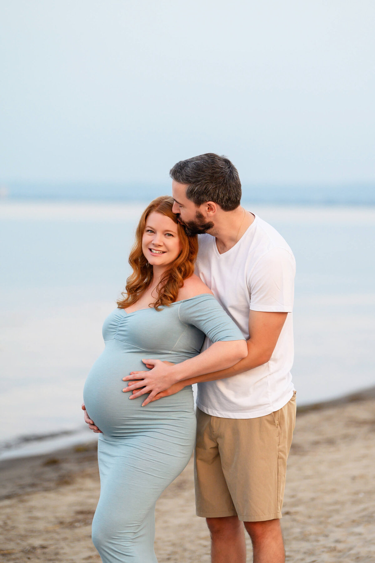 husband and wife cuddling on the beach for their summer maternity photos in Burlington, On