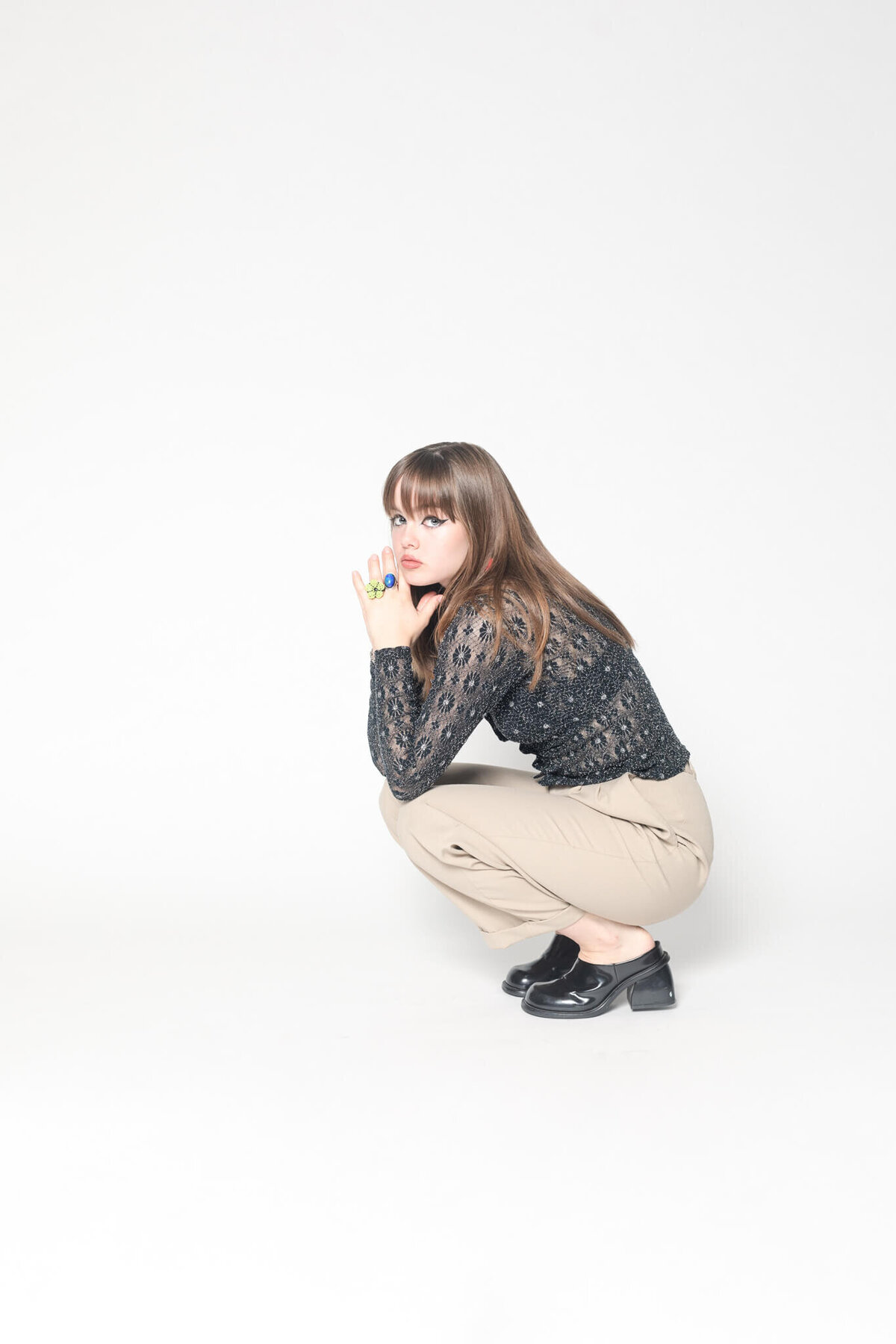 A woman with long brown hair crouches against a white backdrop. She is wearing a black lace top, beige pants, and black shoes. Her hands are together in front of her face.