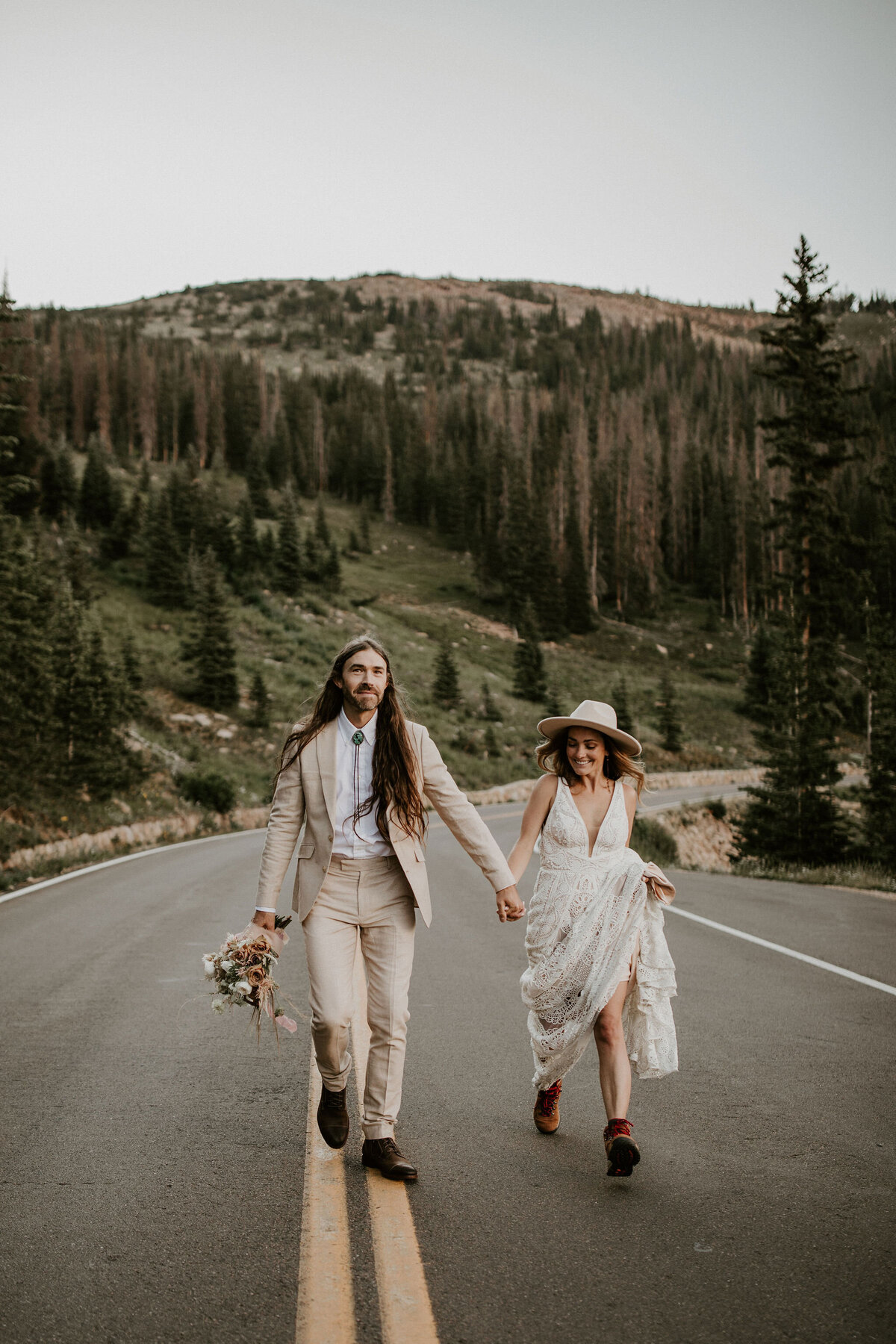 Bride and groom wearing an ivory suit and wedding gown walking on a mountainous road holding hands.
