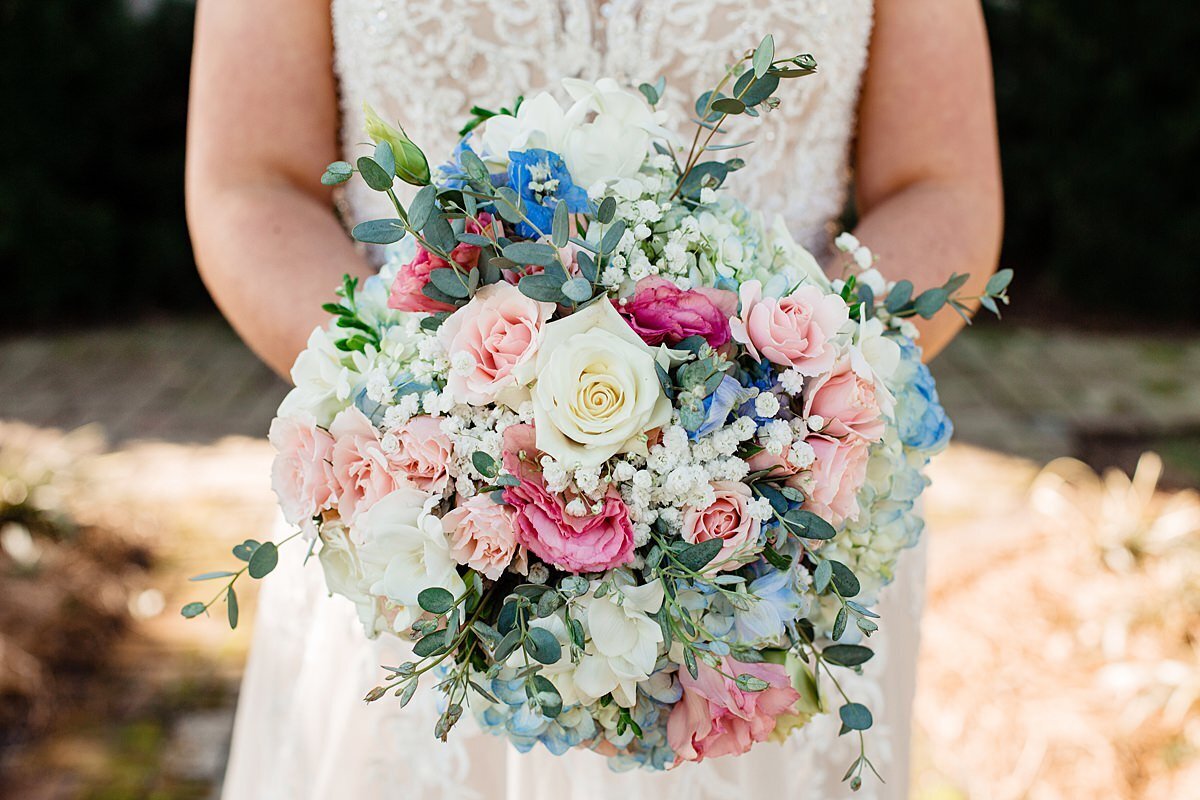 Detail of a peach, pink, ivory, and blue bridal bouquet. The bride holding the bouquet is wearing a sleeveless white lace dress.