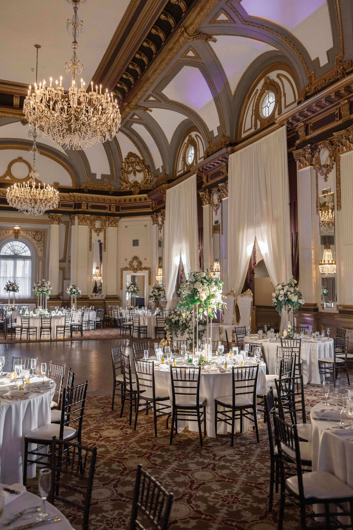 An elegant banquet hall set for an event with round tables covered in white tablecloths, floral centerpieces, and black chairs. Large chandeliers hang from the ornate ceiling, and white drapes adorn the tall windows.