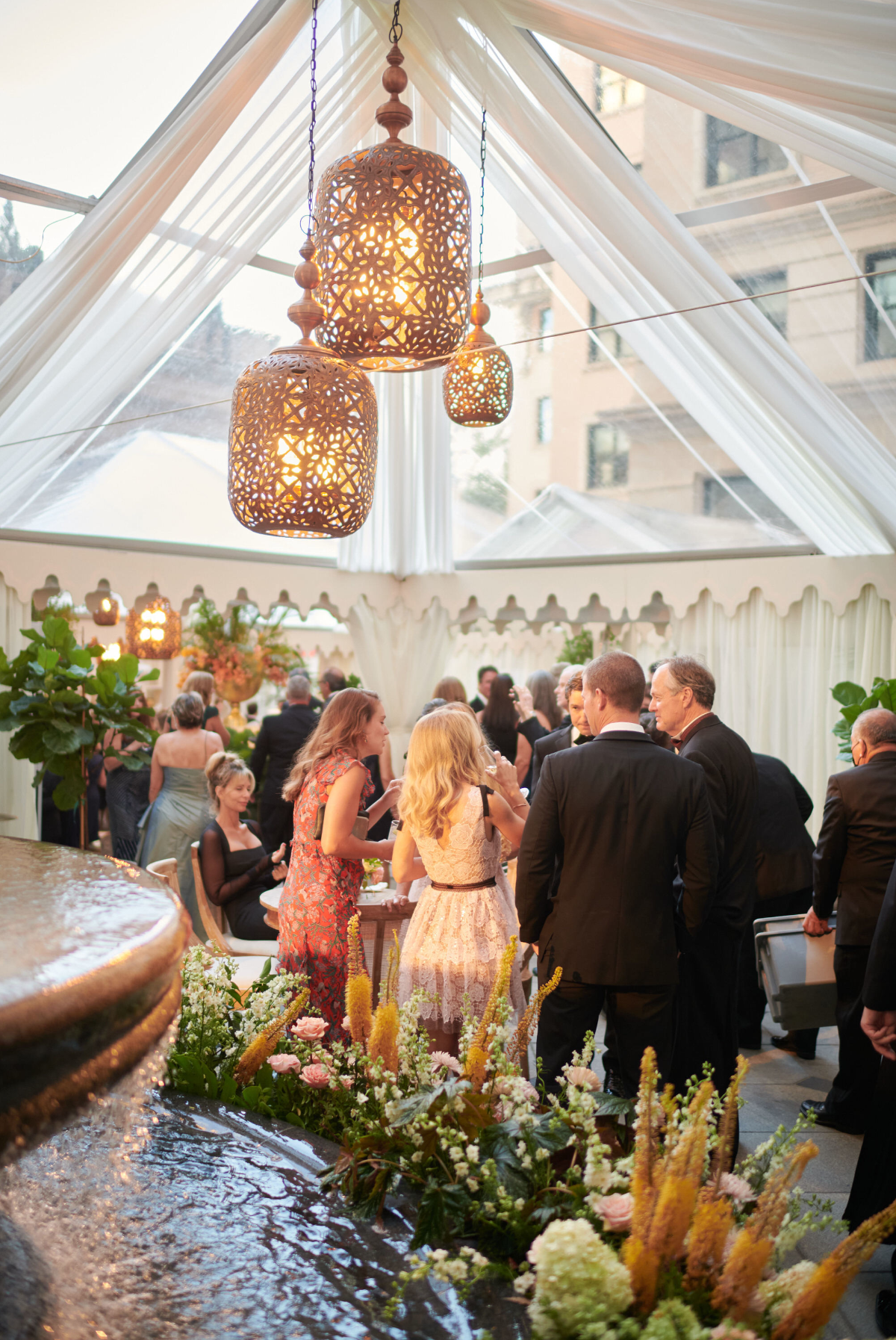This stunning photo was captured at Washington D.C.'s prestigious The Willard Intercontinental, renowned as one of the top wedding venues and hotels in DC. The talented photographer, Shawn Connell, beautifully immortalized a lively indoor wedding reception taking place under a white draped tent adorned with elegant hanging lanterns. As guests, elegantly dressed in formal attire, enjoyed mingling and conversation around tables decorated with gorgeous floral arrangements, a fountain adorned with flowers added a touch of charm in the foreground. Additionally, Clayton Films, Videographers, were also present to capture the special moments of this beautiful event.
